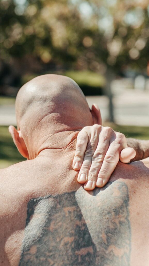 Bald man with tattoo holds sore shoulder outdoors, expressing pain relief.